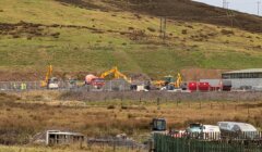 A construction site with excavators, a cement mixer, trucks, and workers set against a grassy hillside.