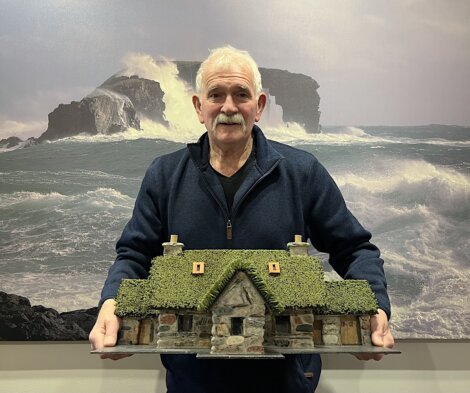 An older man with a mustache holds a detailed model of a stone cottage with a thatched roof. Stormy ocean waves and cliffs are visible in the background image.