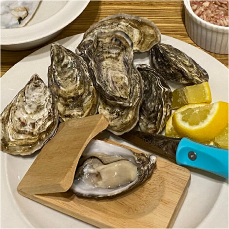 A plate of unopened oysters with a lemon wedge, an oyster shucking knife with a blue handle, and a wooden oyster holder. One oyster is partially opened.