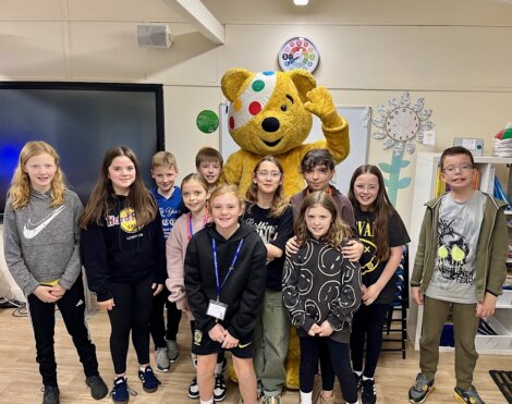 A group of children pose indoors with a large bear mascot in a classroom setting.