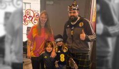 Four people pose indoors; an adult with a crown and kilt gives a thumbs up, another adult smiles beside two children dressed in sports-themed clothing.