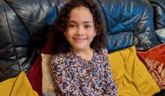 A young girl with curly hair, wearing a floral top, sits on a leather sofa with yellow and red cushions, smiling at the camera.