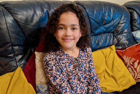 A young girl with curly hair, wearing a floral top, sits on a leather sofa with yellow and red cushions, smiling at the camera.