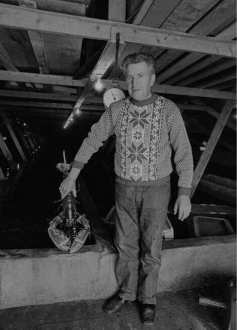 A man wearing a patterned sweater stands in a wooden structure, holding a large lobster with one hand.