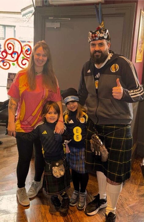 A man, woman, and two children stand indoors; the man and children wear kilts and sports shirts, and the man wears a crown and thumbs up. All are smiling at the camera.
