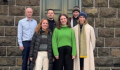 Six people stand together in two rows, posing for a group photo in front of a stone building with a closed door and window.