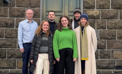 Six people stand together in two rows, posing for a group photo in front of a stone building with a closed door and window.