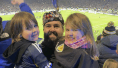 A man wearing a decorated tam o’ shanter and two children smile at the camera in a crowded football stadium.