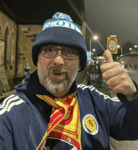 Man wearing a Scotland hat and scarf gives a thumbs-up on a city street at night, with streetlights and people visible in the background.