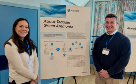 Two people stand smiling next to a presentation board titled "About Tagedale Green Ammonia" at what appears to be an informational event.