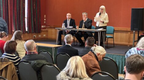 A woman stands at a podium reading from a paper while two men sit at a table on stage facing an audience in a meeting room.