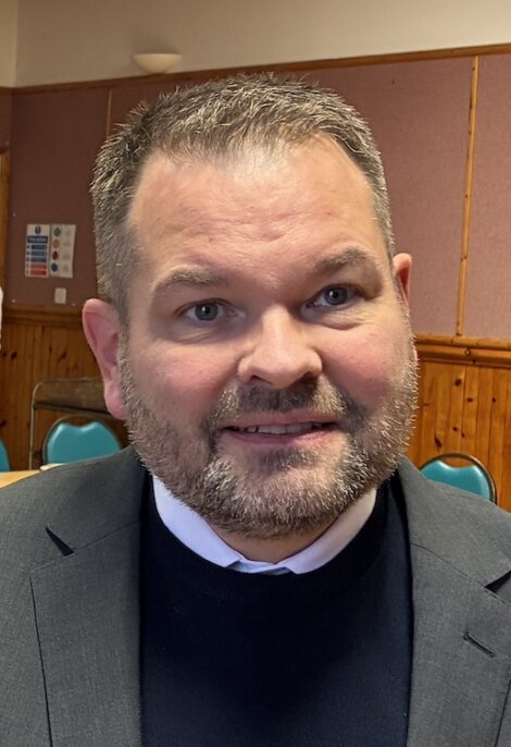 A man with short hair and a beard, wearing a dark suit and sweater, is seated indoors in a room with wood paneling and green chairs.