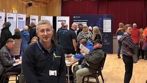 A man with a name badge stands in the foreground at a busy indoor community event with people talking and reading information boards in the background.