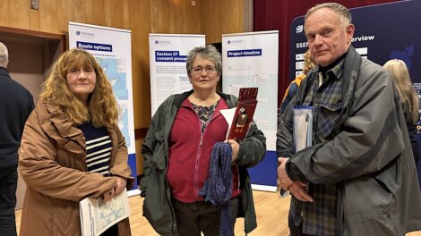 Three adults stand indoors holding documents, with informational display boards about route options, consent process, and project timeline in the background.