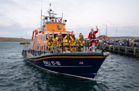 A group in yellow jackets and Santa hats, including one dressed as Santa, stands on an orange RNLI lifeboat approaching a dock with a crowd in the background.