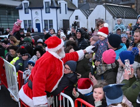 Santa Claus greets and points to a child in a crowd of people behind barriers at an outdoor holiday event.