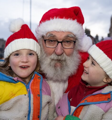 An older man dressed as Santa Claus poses outdoors with two smiling children in winter jackets and red-and-white knit hats.
