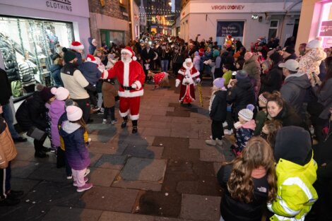 A crowd of people, including children, lines a street as individuals dressed as Santa Claus and Mrs. Claus walk through during a festive event.