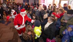 Santa Claus greets a crowd of adults and children on a decorated street at night; people are bundled up in winter clothing and there is a small brass band in the background.