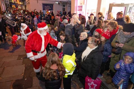 Santa Claus greets a crowd of adults and children on a decorated street at night; people are bundled up in winter clothing and there is a small brass band in the background.