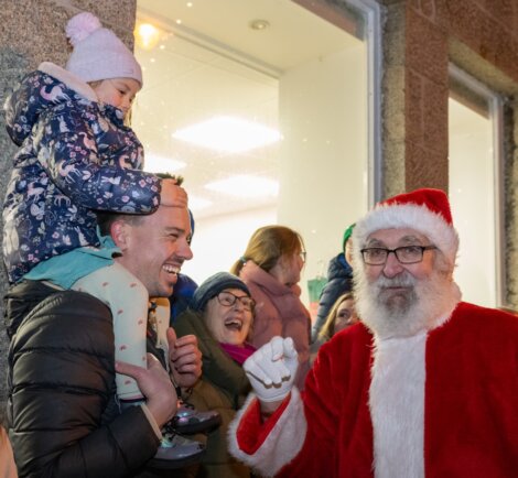A man carries a child on his shoulders while smiling at a person dressed as Santa Claus; several people stand and watch in the background.