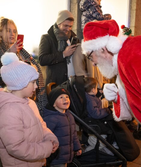 A man dressed as Santa Claus leans down to speak with two young children, one in a stroller, while adults watch and take photos.