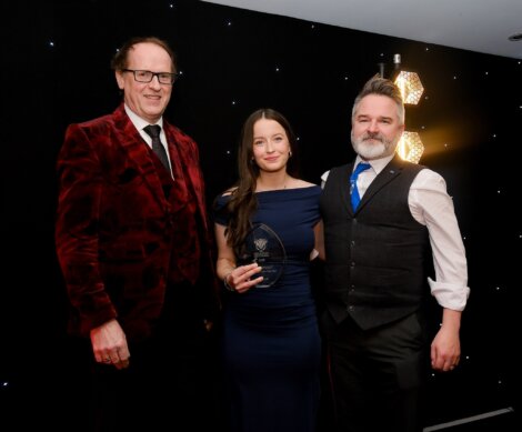 Three people pose indoors; the woman in the center holds an award. All are dressed formally, standing in front of a black backdrop with small lights.