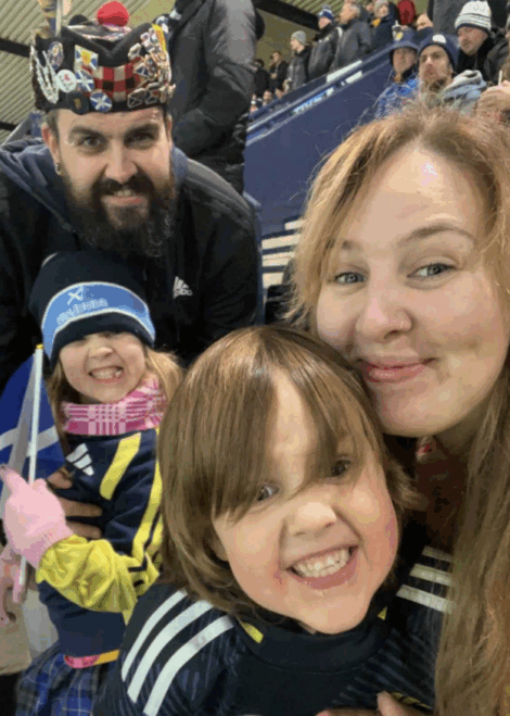 A man, woman, and two children smile for a selfie at a stadium event, with fans and seats visible in the background.