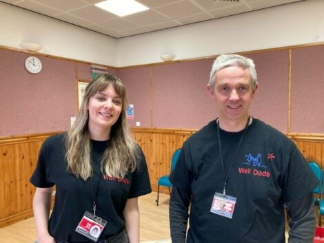 Two people wearing "Well Dads" t-shirts and name badges stand indoors in a community room with wooden paneling and noticeboards in the background.