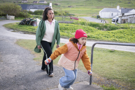 A woman in a green coat walks behind a young girl in a pink jacket and red hat who is running on a rural path with houses and green fields in the background.