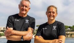 Two people in black team shirts stand side by side with arms crossed, smiling outdoors near water on a sunny day.