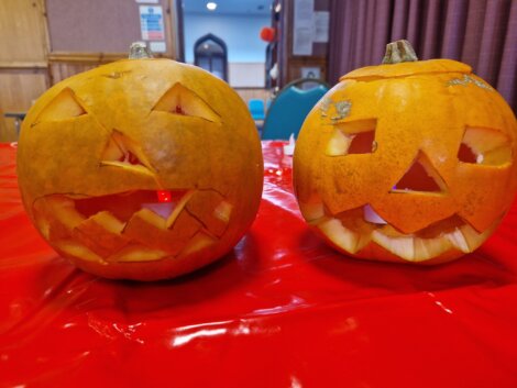 Two carved pumpkins with triangle eyes and jagged mouths are displayed on a red tablecloth in a room with wood-paneled walls and chairs in the background.