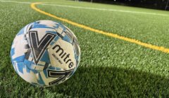 A Mitre soccer ball rests on artificial turf near a yellow line at an outdoor field under nighttime lighting.