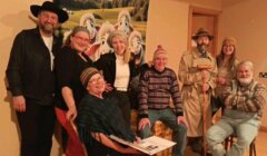 A group of seven adults in costume pose indoors; some are standing, some are seated, and a backdrop with cartoon dogs is behind them.