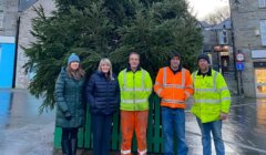 Five people stand in front of a large outdoor Christmas tree, with some wearing high-visibility jackets and wet pavement beneath them.