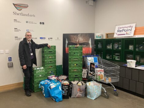 A man stands beside stacked green crates, shopping bags, and a shopping cart filled with items in front of a wall with Shetland Arts and Community RePaint signage.