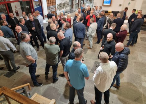 A large group of men stand and converse in a spacious indoor area, some holding drinks, suggesting a social or networking event.