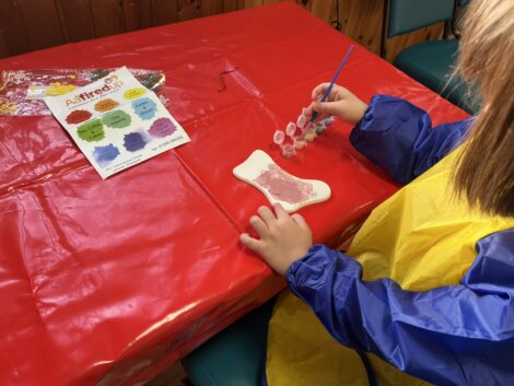 A child wearing a yellow and blue smock paints a ceramic stocking with pink paint at a table covered with a red plastic tablecloth. An instruction sheet and paint set are nearby.