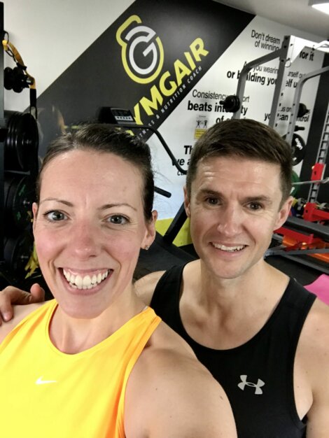 Two people in athletic wear smile for a selfie at a gym with motivational quotes on the wall and fitness equipment in the background.
