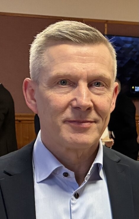 A middle-aged man with short gray hair in a blue collared shirt and dark blazer stands indoors, looking at the camera.
