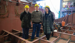 Three people wearing hard hats and winter clothing stand inside a metal structure under construction in an industrial workshop.