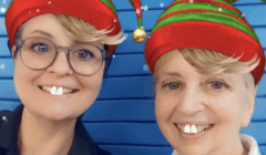 Two people with glasses and elf hats smile at the camera. A filter adds exaggerated front teeth, freckles, and short blond hair to their faces. Blue background.
