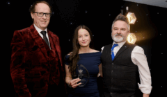 Three people in formal attire stand together at an event; the woman in the center holds a transparent award and all are looking at the camera.