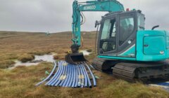 A blue excavator is parked on grassy terrain next to a stack of blue pipes, with waterlogged ground visible in the background.