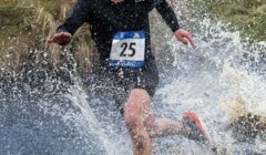 A runner wearing a black outfit and bib number 25 splashes through a muddy water obstacle during a cross-country race.