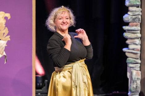A woman in a gold crown and sash stands on stage, smiling and making a heart shape with her hands.