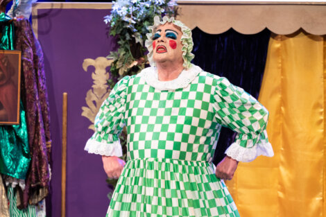A performer in theatrical makeup and a green-and-white checkered dress stands with hands on hips on stage, wearing a frilly bonnet.