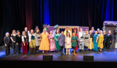 A group of adults and children in colorful costumes stand on a theater stage, posing for a group photo in front of stage props and curtains.