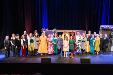 A group of adults and children in colorful costumes stand on a theater stage, posing for a group photo in front of stage props and curtains.
