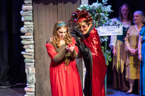 A woman in a red dress holds an apple while another person in a red cape and mask gestures toward her on stage, with onlookers and a sign in the background.